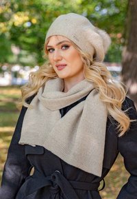 Un bonnet en tricot beige avec un pompon et une écharpe assortie drapée autour du cou, porté par-dessus un manteau sombre. Les matériaux semblent doux et texturés.