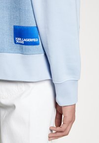 Felpa azzurro chiaro con pannello in denim e etichetta blu con la scritta "KARL LAGERFELD JEANS." Tessuto liscio, polsini a coste.