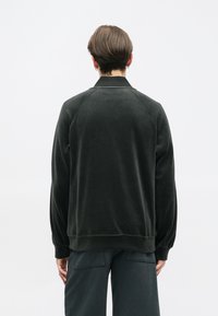 Sweatshirt de veludo suave em verde escuro, com um colarinho e punhos canelados, apresentando um corte descontraído e mangas raglan. Design minimalista, sem padrões visíveis.