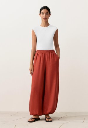 Woman wearing a white sleeveless top and loose-fitting rust-colored pants with an elastic waist, standing with one hand in pocket and black sandals.