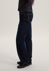 Jeans di denim blu scuro con taglio dritto, caratterizzati da cuciture bianche, tasche posteriori e una texture liscia. Indossati con calzature nere.