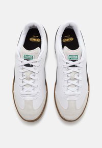 Witte sneakers met een soepel leren bovenwerk, suede accenten, zwart en gouden branding, en een gestructureerde gumrubberen zool. Vetersluiting.