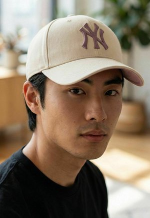 Junger Mann mit kurzen schwarzen Haaren, trägt eine beige Baseballkappe mit dunklem Logo und ein schwarzes Hemd, in einem sanft beleuchteten Innenraum.