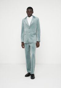 Svečano odijelo od baršuna u svijetloplavoj boji, s revnim ovratnikom i dva prednja džepa, upareno s bijelom košuljom s ovratnikom i crnim cipelama.