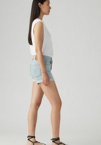 Top blanco sin mangas, pantalones cortos de mezclilla azul claro con dobladillos enrollados, y sandalias negras con tiras. Vista lateral contra un fondo neutro.
