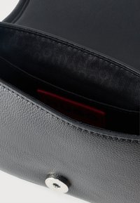 Μαύρη τσάντα από δερματίνη με υφή, μερικώς ανοιχτή που δείχνει εσωτερική επένδυση από λείο μαύρο ύφασμα και κόκκινη ετικέτα στο εσωτερικό. Ορατό ασημένιο μαγνητικό κλείσιμο.