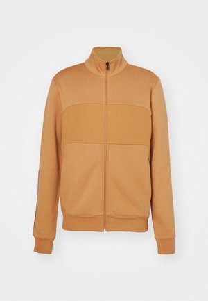 Lys brun zip-up jakke med høy krage, som har et panelert design på brystet, ribbede mansjetter og en glatt, myk tekstur.