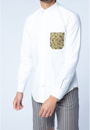 Weißes Hemd mit Knopfleiste, khakifarbenem Camouflage-Taschenmotiv, langen Ärmeln und geradem Schnitt. Kombiniert mit grauen, gestreiften Shorts.