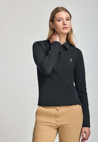 Camisa polo negra de manga larga con cuello, que presenta una tapeta de botones y un pequeño logotipo blanco en el pecho; combinada con pantalones beige.