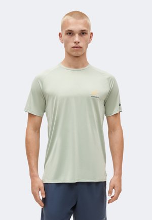 Młody mężczyzna z krótkimi blond włosami, ubrany w jasnozieloną koszulkę Nike Dri-FIT i ciemne spodenki, stoi na tle jednolitego jasnego tła.