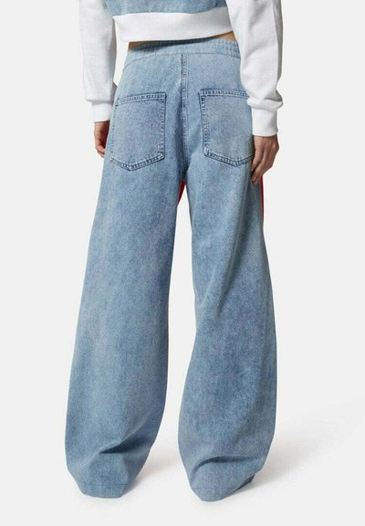 Jeans in denim baggy blu chiaro con vita alta, dotati di grandi tasche posteriori e una texture liscia. Accenti rossi sui lati visibili.