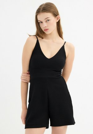 Mono negro texturizado con escote en V, tirantes finos y cintura ceñida. Los pantalones cortos llegan hasta la mitad del muslo, mostrando un acabado suave.