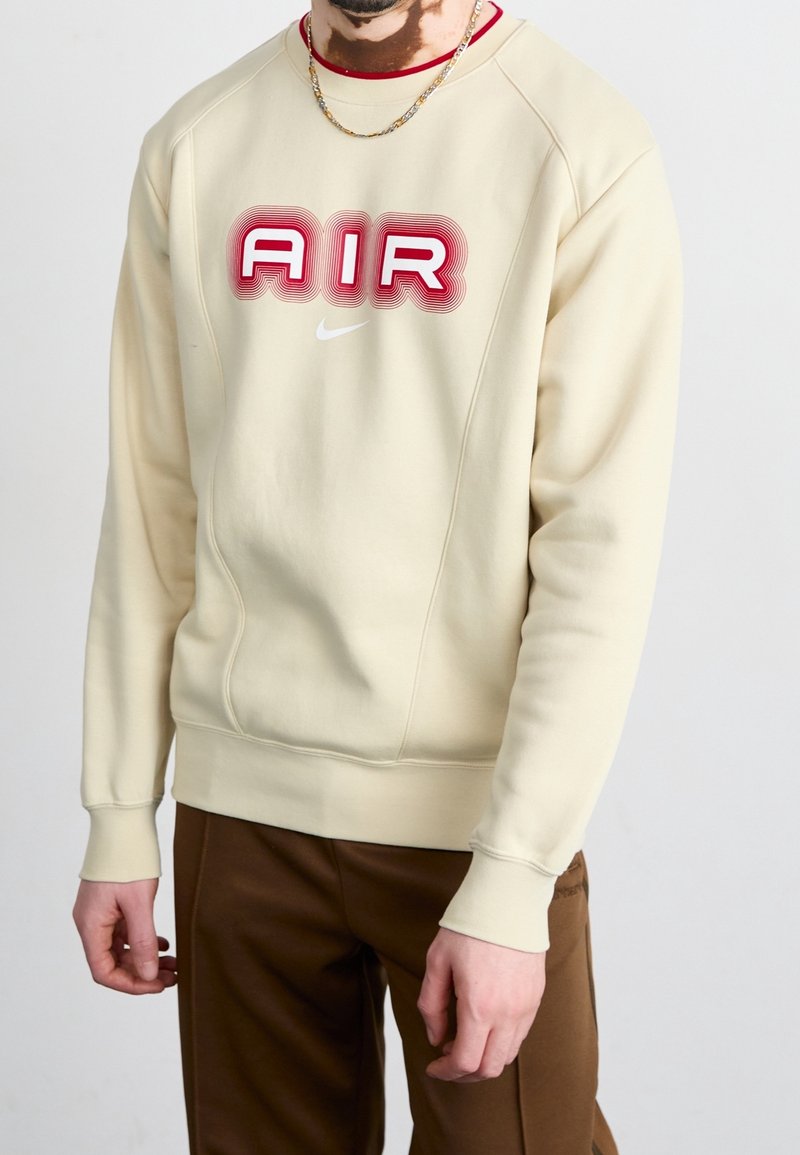 Personne portant un sweat beige avec un texte rouge "AIR" et un logo Nike blanc, associé à un pantalon marron et un collier chaîne en or.