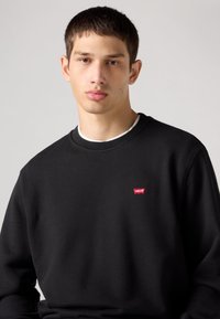 Crna majica s kapuljačom od pamučnog runa s okruglim izrezom i rebrastim manšetama. Na prsima ima mali crveni Levi's logo.