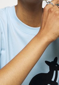 Camiseta de algodón de color azul claro con un gráfico de la silueta de un gato negro. Anillos plateados visibles en los dedos y piel texturada. Cuello redondo simple.
