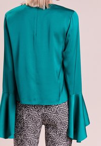 Turkusowa satynowa bluzka z rozszerzanymi rękawami, z detalem na szwie tylnym, w połączeniu z czarno-białymi spodniami w wzory, charakteryzującymi się dopasowanym krojem.