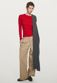 Rotes Langarmoberteil, beigen weiten Hosen und einen dunkelgrauen Pullover, der über eine Schulter hängt. Die Schuhe sind dunkelbraune Loafer.
