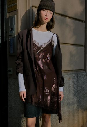 Jonge vrouw met een donkere muts, een wit shirt met lange mouwen onder een bruin bloemenjurkje, en een oversized donker colbert, staand tegen een muur.