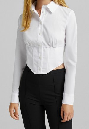 Chemise blanche à manches longues avec une taille cintrée, des fermetures à boutons et un col pointu. Présente des découpes latérales et des fronces au niveau de la poitrine.