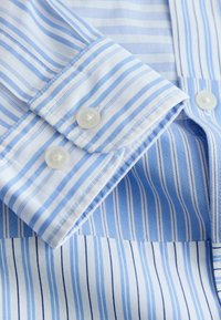 Poignet de chemise à manches longues rayée bleu clair et blanc avec deux boutons blancs et des coutures détaillées sur tissu en coton.