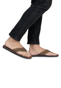 Tongs marron avec une sangle en faux cuir texturé et une semelle en caoutchouc blanche. Pied positionné sur un jean sombre, présentant un design lisse et minimal.