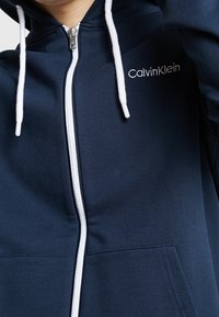 Sudadera con cremallera de color azul marino, hecha de tejido suave, con una cremallera blanca, cordones y un bolsillo delantero. Logotipo "Calvin Klein" en el lado izquierdo del pecho.