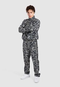 Camouflagetrainingspak in grijs en zwart, gemaakt van gestructureerde stof. Voorzien van een jas met rits en opstaande kraag en een broek met elastische tailleband.