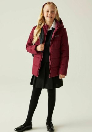 Niña con el cabello rubio largo que lleva una chaqueta acolchada burdeos, uniforme escolar negro, medias negras, zapatos negros y lleva una mochila negra.
