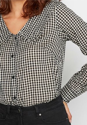 Kobieta w czarno-białej koszuli w kratę z falbaną przy kołnierzu i czarnych spodniach, ręka oparta na biodrze na jednolitym tle.