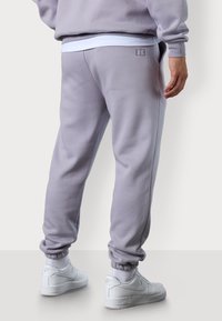 Hellgraue Sweatpants mit Rippbündchen und einem gestickten Logo auf der Rückseite. Kombiniert mit weißen Sneakers, was einen lässigen Look ergibt.