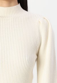 Kremowy, prążkowany sweter dzianinowy z wysokim kołnierzem i bufkami na ramionach, o miękkiej fakturze i dopasowanym kroju.