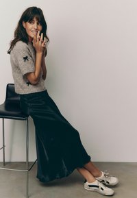 Maglione a maniche corte in grigio chiaro con dettagli a fiocco nero, abbinato a una gonna fluida in raso nero e sneakers beige con dettagli neri.