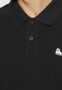Polo noir à col avec deux boutons noirs de marque et une petite écusson de chaussure blanche sur la poitrine droite.