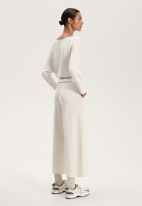 Világos bézs színű nappali szett, amely egy hosszú ujjú felsőből áll, széles nyakkivágással, és ellazult szabású nadrágból zsebekkel, sportcipőkkel párosítva.