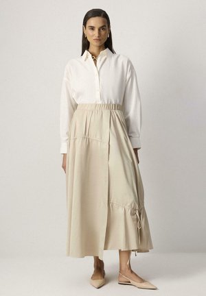 Žena v bielej košeli zapínanej na gombíky, zastrčenej do béžovej midi sukne s elastickým pásom a záhybmi, v béžových špicatých balerínach.