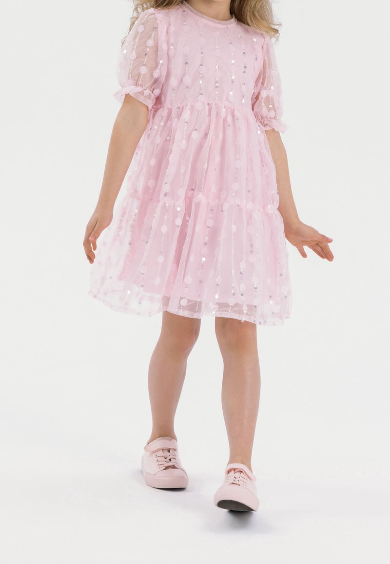 Robe rose en tissu transparent texturé avec manches bouffantes et jupe à volants, ornée de sequins réfléchissants. Associée à des chaussures roses.
