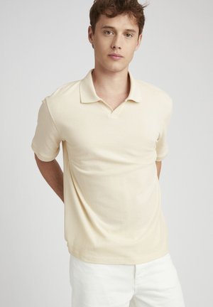 Polo beige en tissu texturé, à manches courtes, avec un col classique et des poignets subtils, associé à un pantalon blanc.