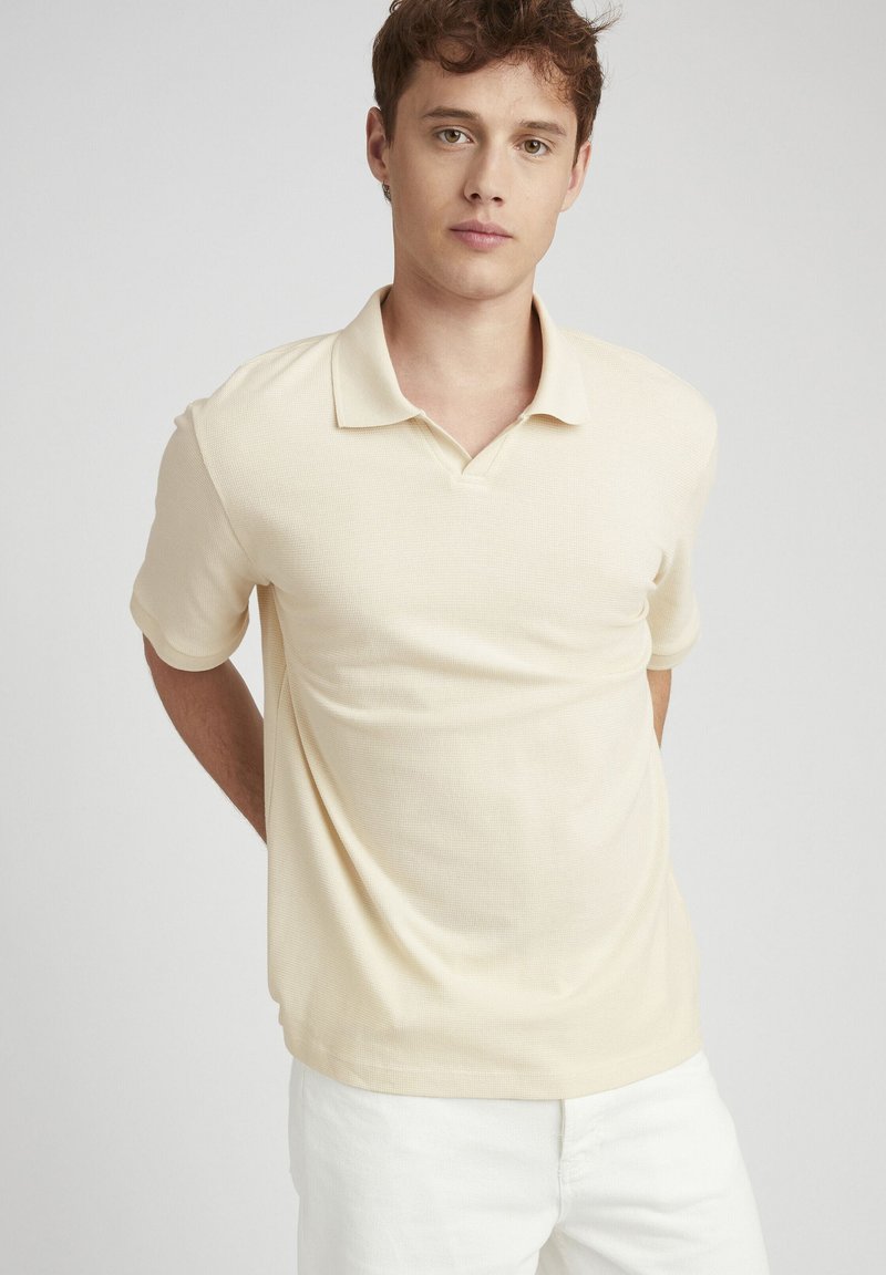 Polo beige en tissu texturé, à manches courtes, avec un col classique et des poignets subtils, associé à un pantalon blanc.