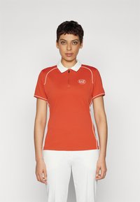 Raudonas polo marškinėliai su balta apykakle ir dekoratyvinėmis juostelėmis, turintys pusiau užtrauktuką ir logotipą ant krūtinės. Lygus, tampres audinys.