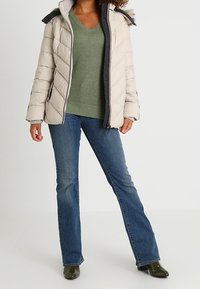 Beżowa pikowana kurtka z kapturem, szarym podbiciem i sztucznym futrem, noszona na zielonym swetrze i niebieskich dzwonach, uzupełniona zielonymi butami.