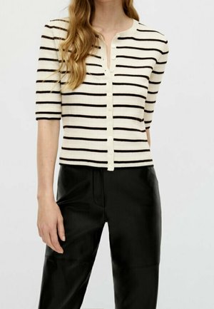 Woman wearing a short-sleeve, buttoned, cream top with black horizontal stripes and black high-waisted pants against a plain background.