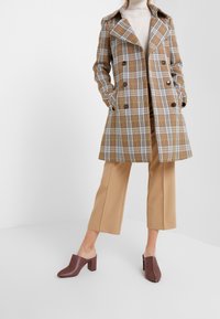 Trench-coat en tartan marron avec boutons double boutonnage, associé à un pantalon cropped beige et des mules à talons bordeaux. Tissu lisse et coupe ajustée.