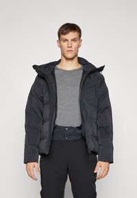 Jachetă puffer neagră cu un model texturat în spirală, cu fermoar în față, glugă ajustabilă și izolație groasă, purtată peste un tricou gri și pantaloni negri.