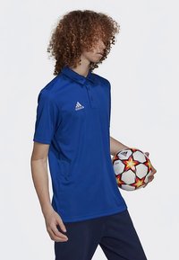 Polo bleu avec col, manches courtes et logo Adidas. Le mannequin tient un ballon de football composé de panneaux noirs et blancs avec des accents rouges.