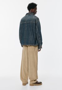 Džínová bunda v tmavo modrej farbe s voľným strihom, dlhými rukávmi a jemným vyblednutím. Skombinovaná s béžovými kordovým nohavicami a hnedou obuvou.