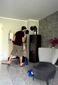 Άνδρας με σορτς και σανδάλια κρεμάει ένα σακάκι σε μια ξύλινη κρεμάστρα σε μοντέρνο σαλόνι με μαύρο καλοριφέρ και γκρι πολυθρόνα.