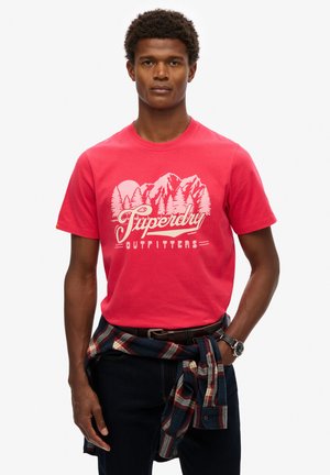 Czerwona bawełniana koszulka z nadrukiem gór i drzew, z napisem "Superdry Outfitters"; zestawiona z ciemnymi dżinsami i związanym w talii flanelowym płaszczem w kratkę.