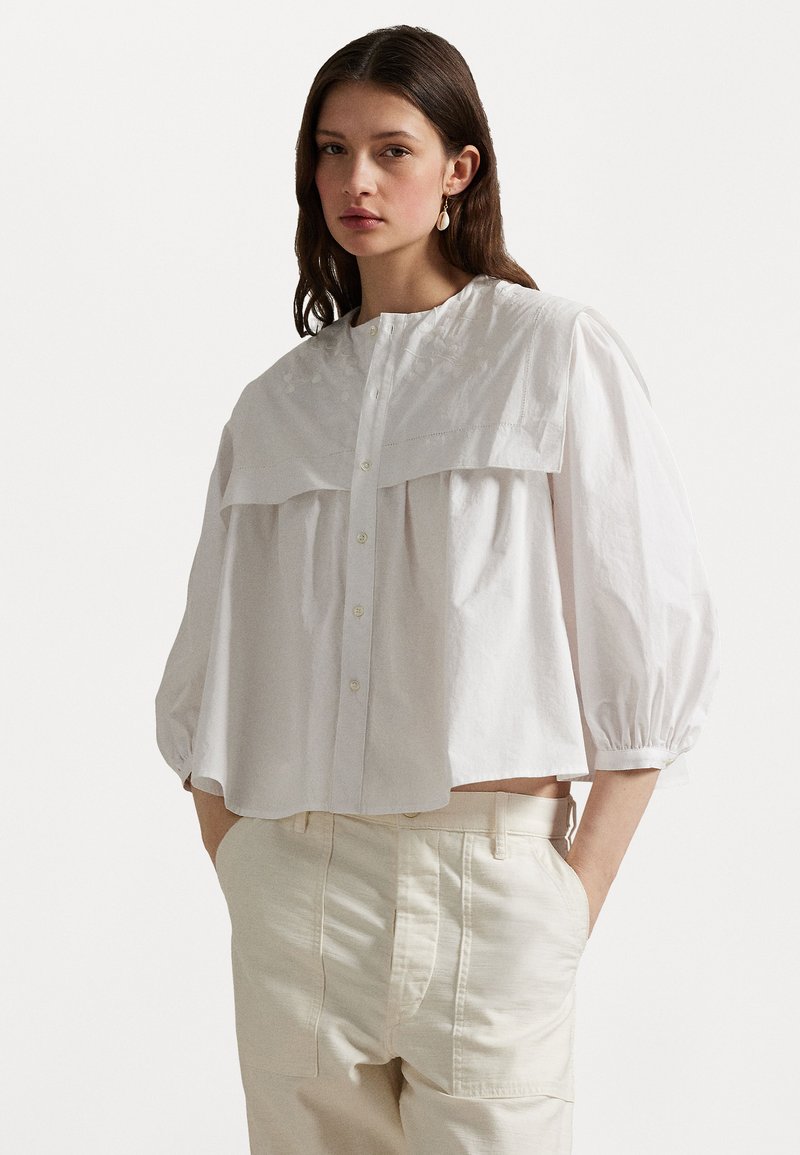 White blouse with puffed sleeves, oversized collar, and front button closure. Smooth cotton fabric with subtle textured details.