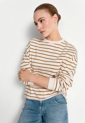 Junge Frau mit heller Haut und zurückgebundenem Haar, die ein weißes und braunes gestreiftes Langarmshirt und eine blaue Jeans trägt, steht vor einer weißen Wand.