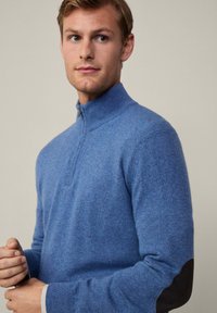 Pullover blu con zip, texture a maglia e toppe in pelle nera sui gomiti. Disponibile in design aderente.